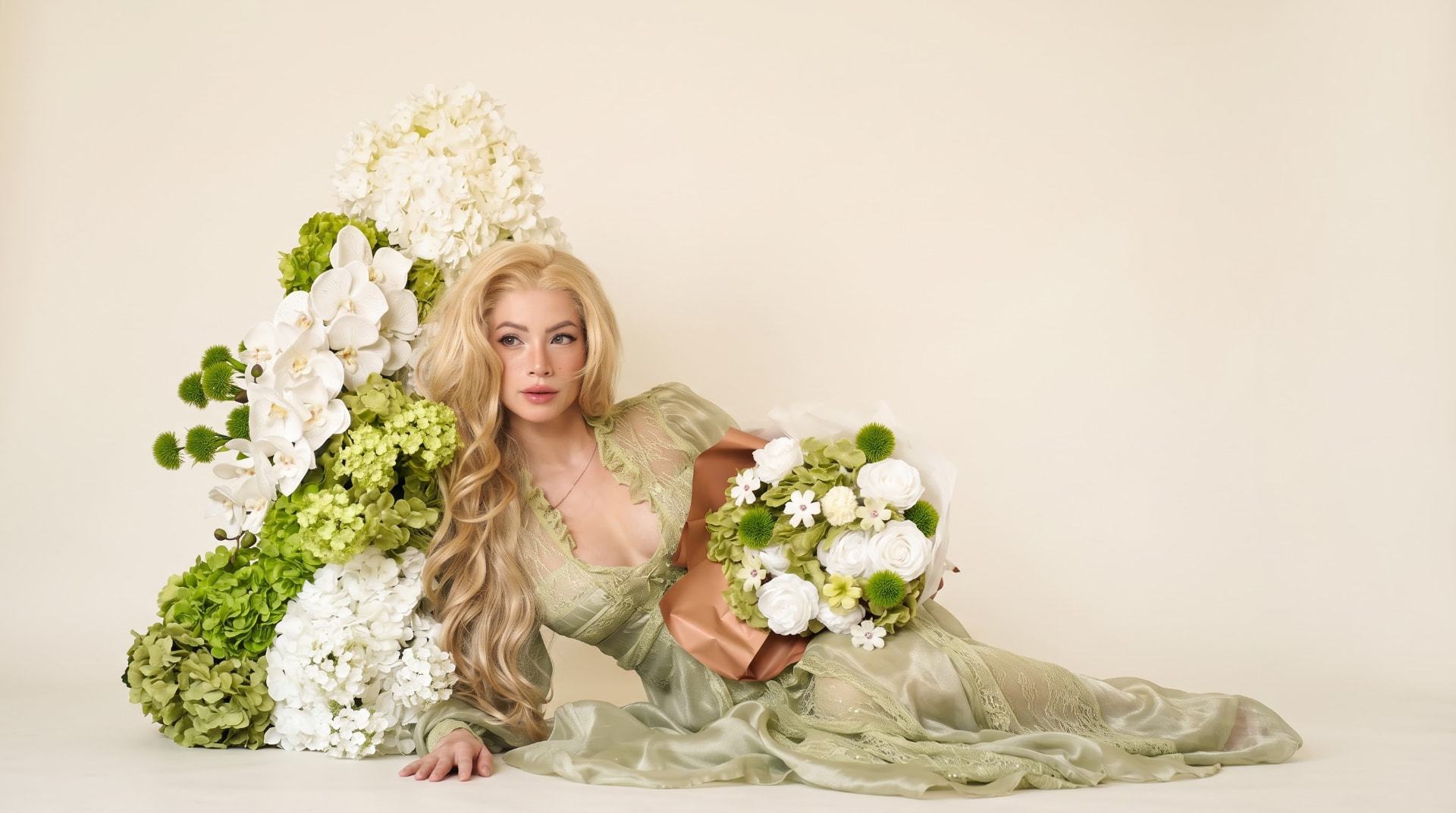 Model in a sage-green lace gown reclining among white and green hydrangea and orchid arrangements, holding a wrapped Fleurmore bouquet